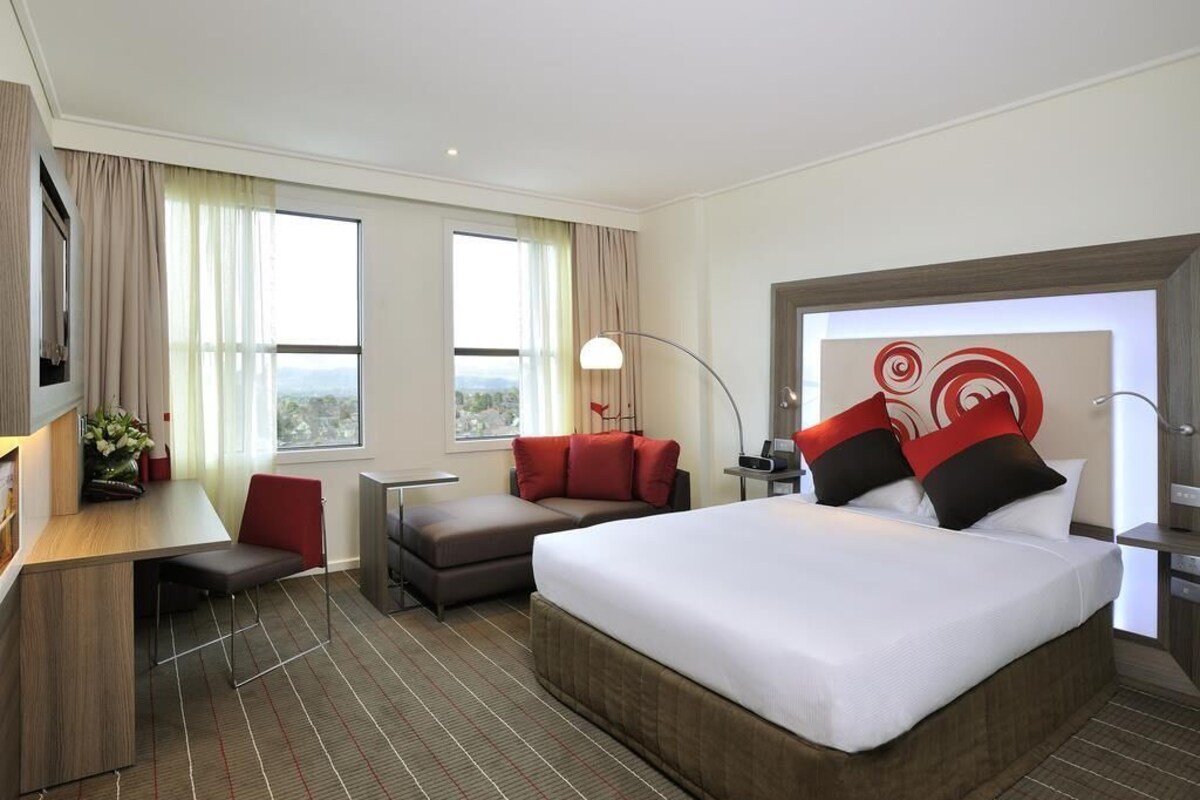 Une chambre d’hôtel avec un grand lit, un bureau, une chaise et un canapé. Il y a une fenêtre avec des rideaux, une lampe et un élément décoratif mural.