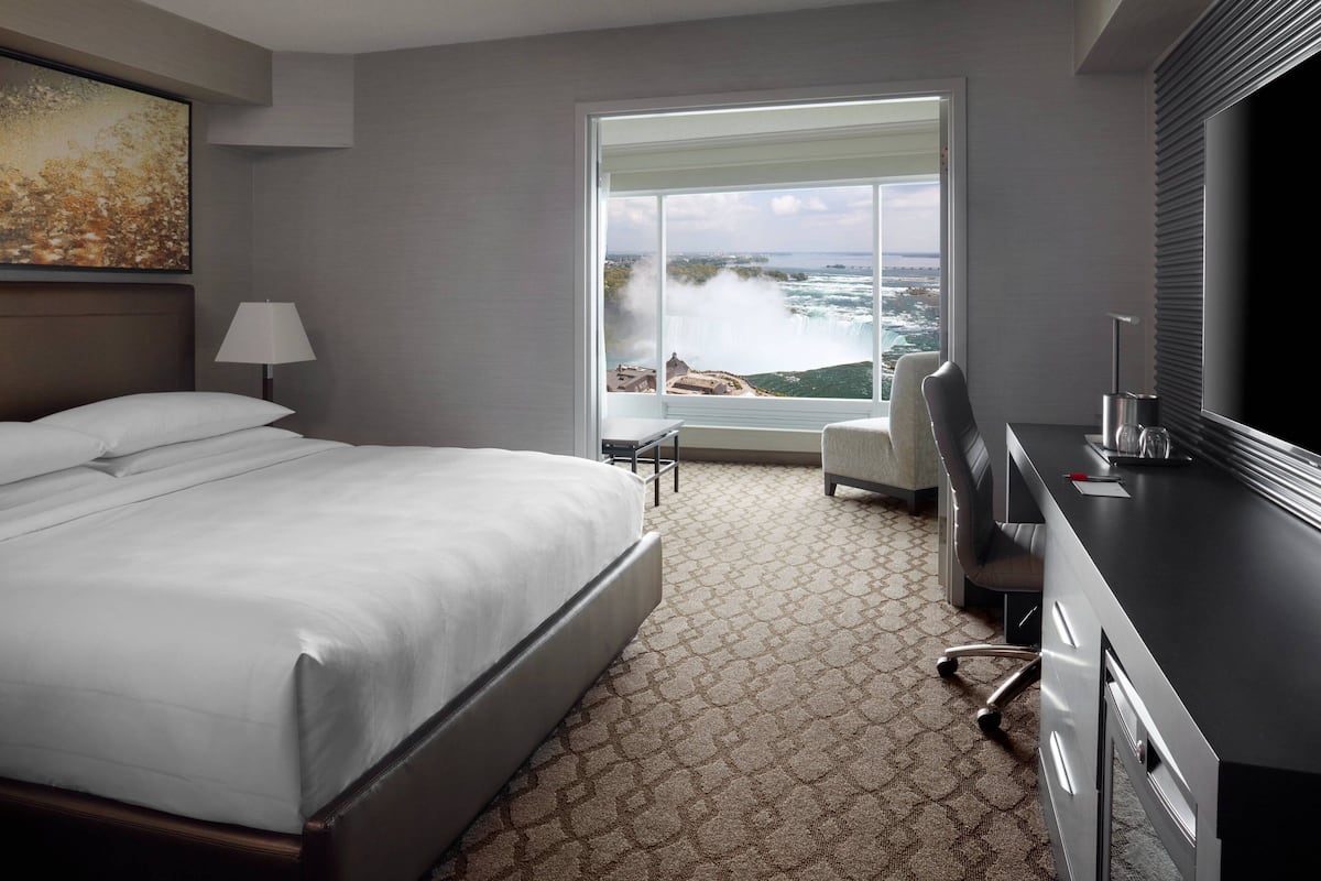 A hotel room with a large bed, a desk, a chair, a TV, and a window with a view of a waterfall.