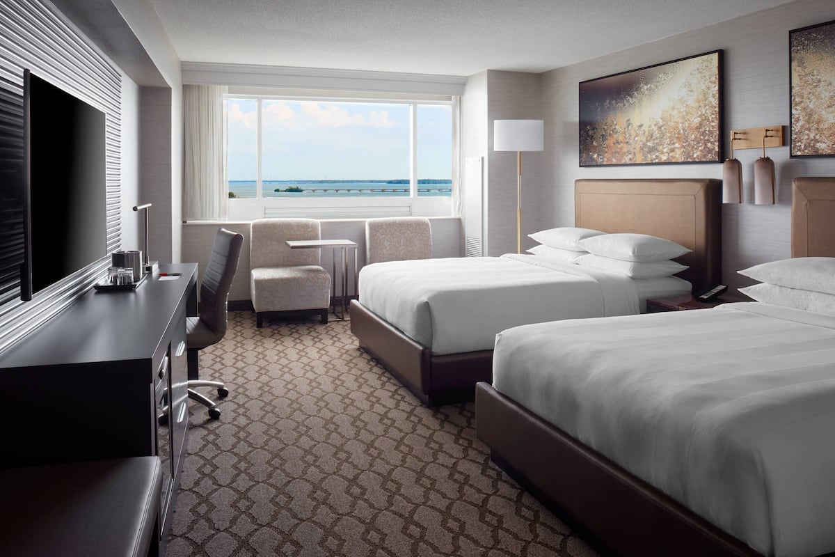 A hotel room with two beds, a desk, a chair, a TV, and a window with a view of the sea.