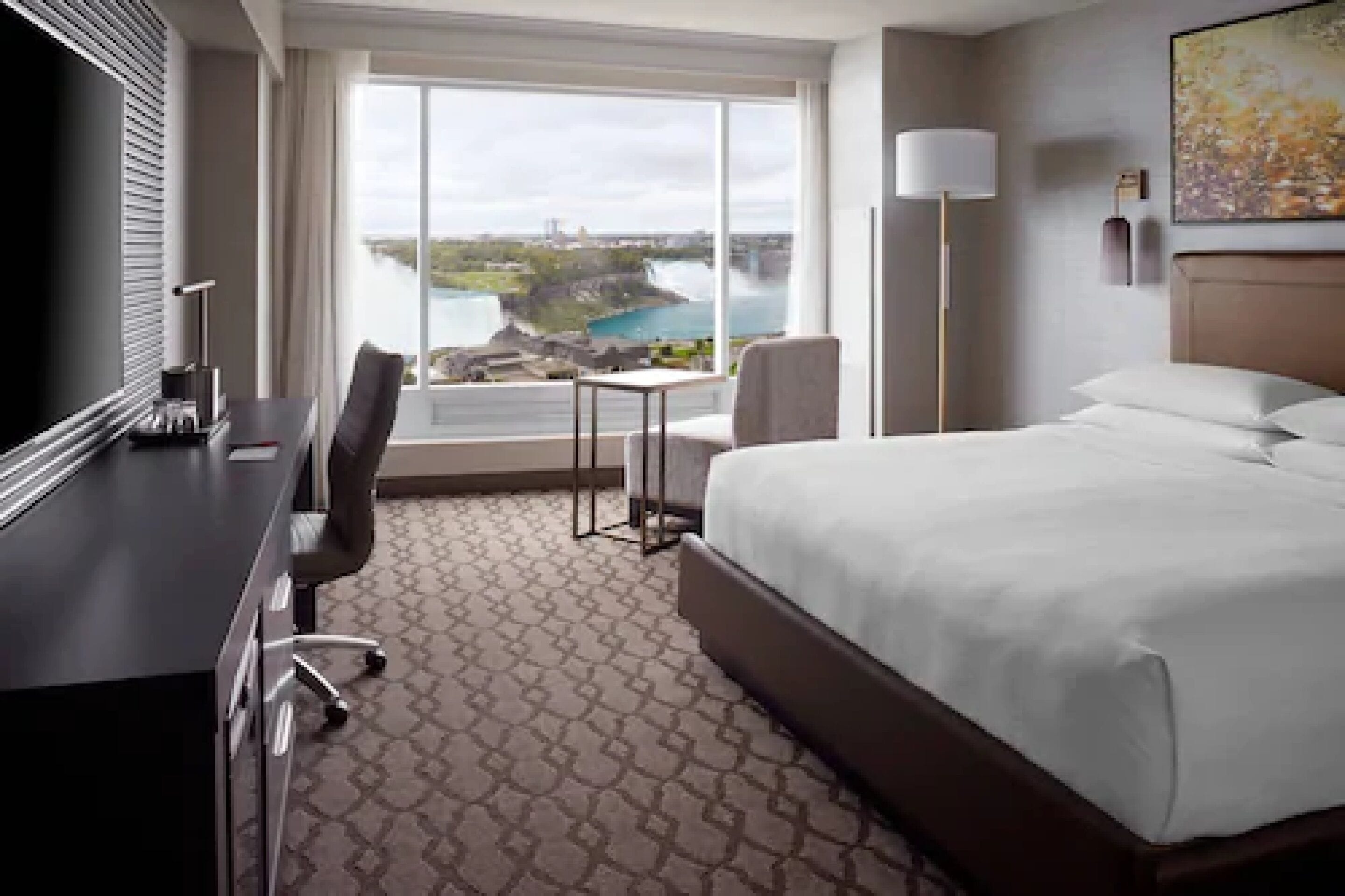 A hotel room with a large bed, a desk, and a chair. There is a window with a view of a body of water and a building.