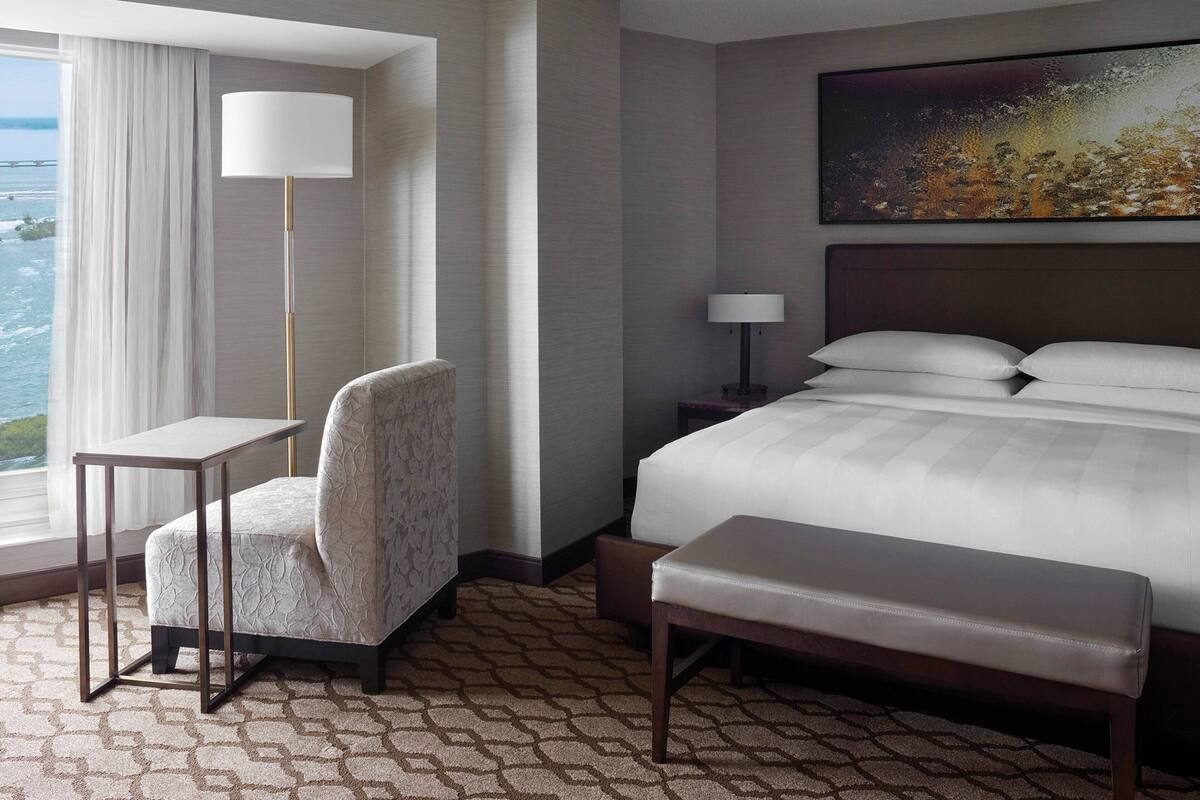 A hotel room with a bed, a desk, a chair, a bench, a nightstand, a lamp, and a painting on the wall.