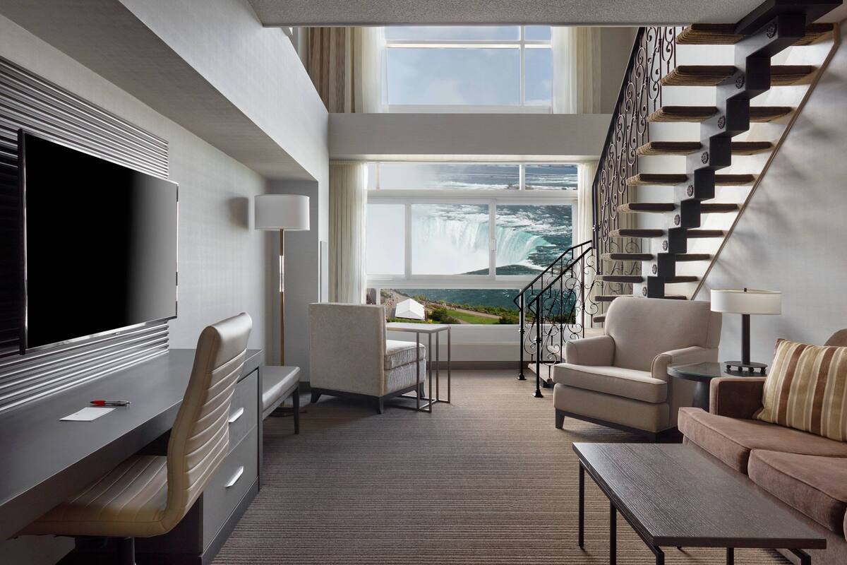 A modern hotel room with a large flat-screen TV, a comfortable sofa, a desk with a chair, and a view of a waterfall through large windows.