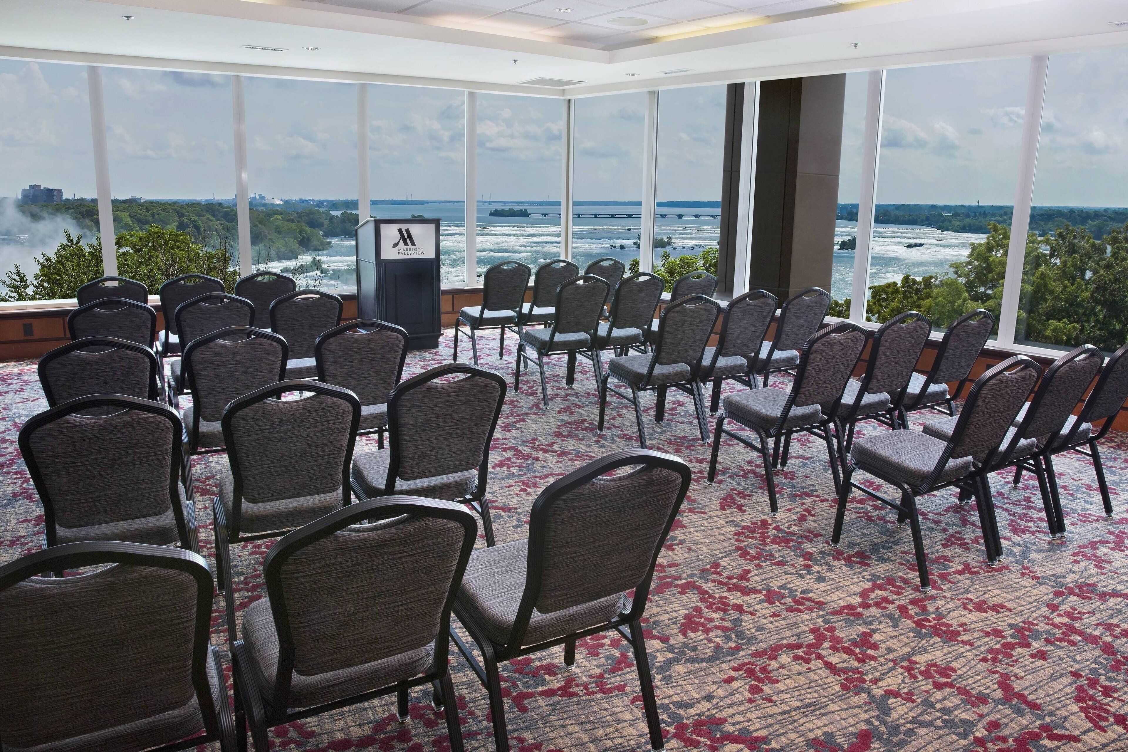 Un salón de conferencias con filas de sillas orientado a un amplio ventanal con vistas a un río y un paisaje urbano.