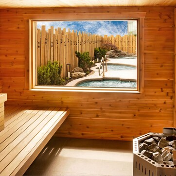 A wooden sauna room with a bench, towels, a window view of a pool and garden, and a stone basin.