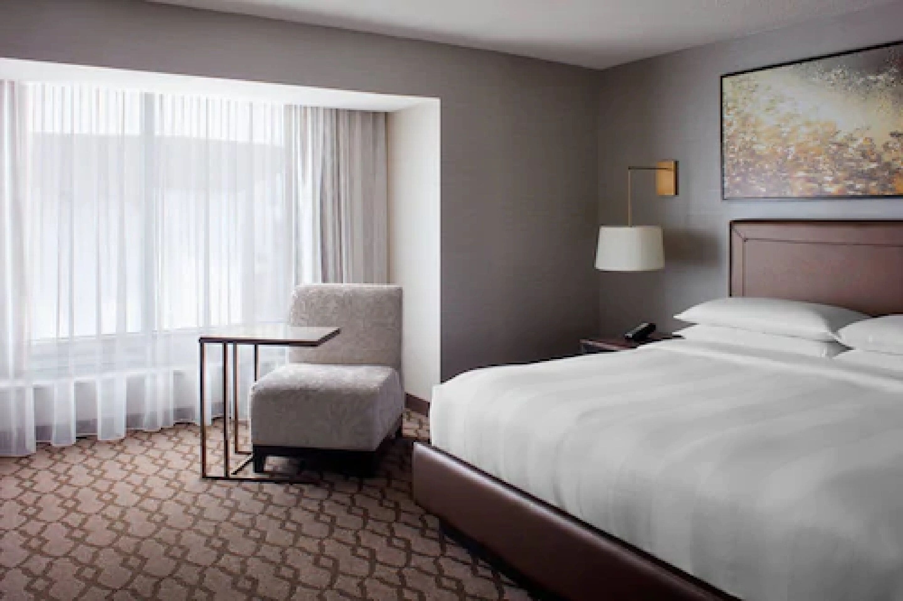 Habitación de hotel con una cama grande, una mesita, una silla, una lámpara y un cuadro en la pared.