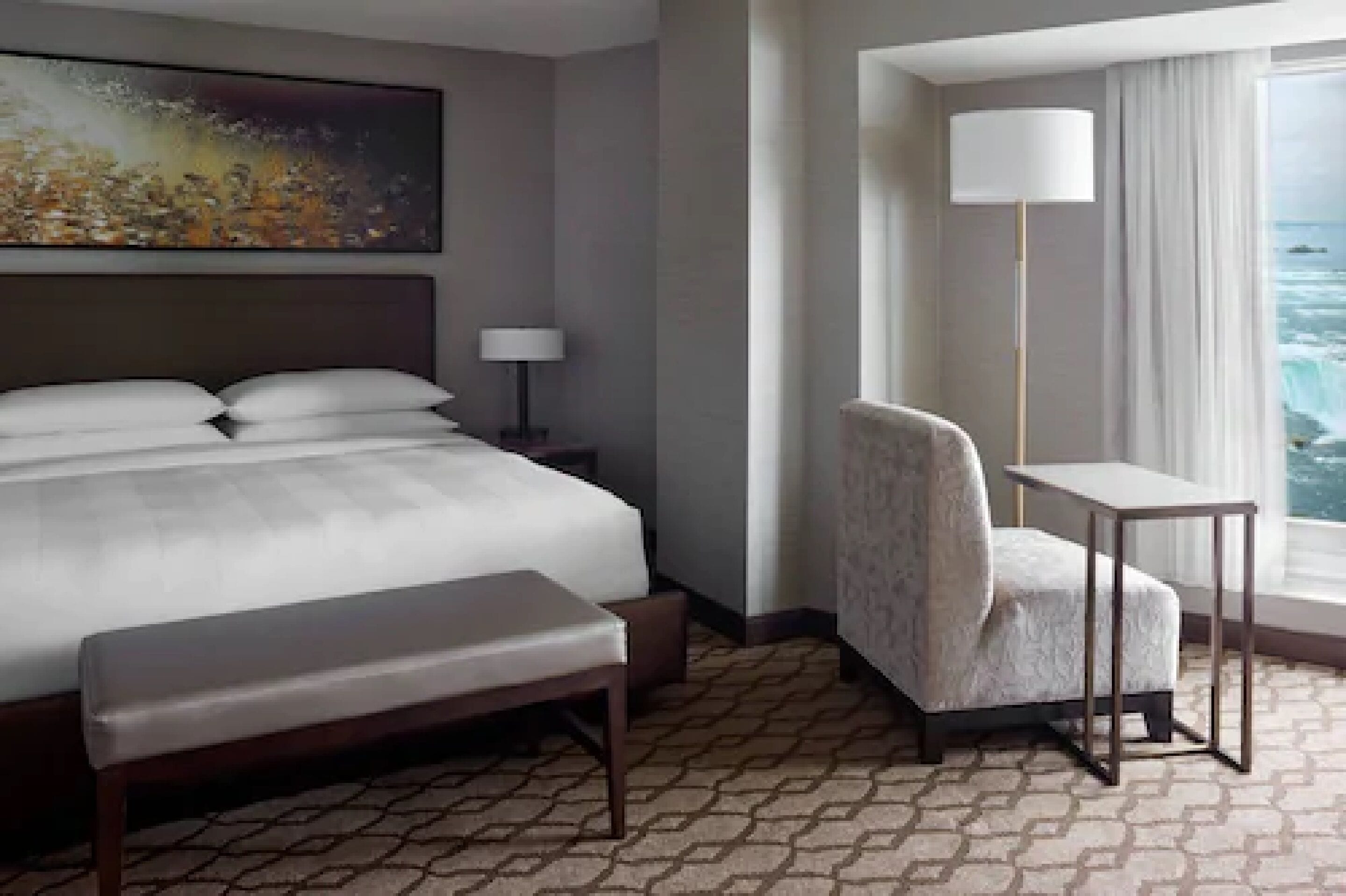 A hotel room with a bed, a bench, a chair, a desk, a lamp, and a painting on the wall.