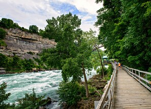 A wooden boardwalk along a river with a rocky cliff and dense foliage.
