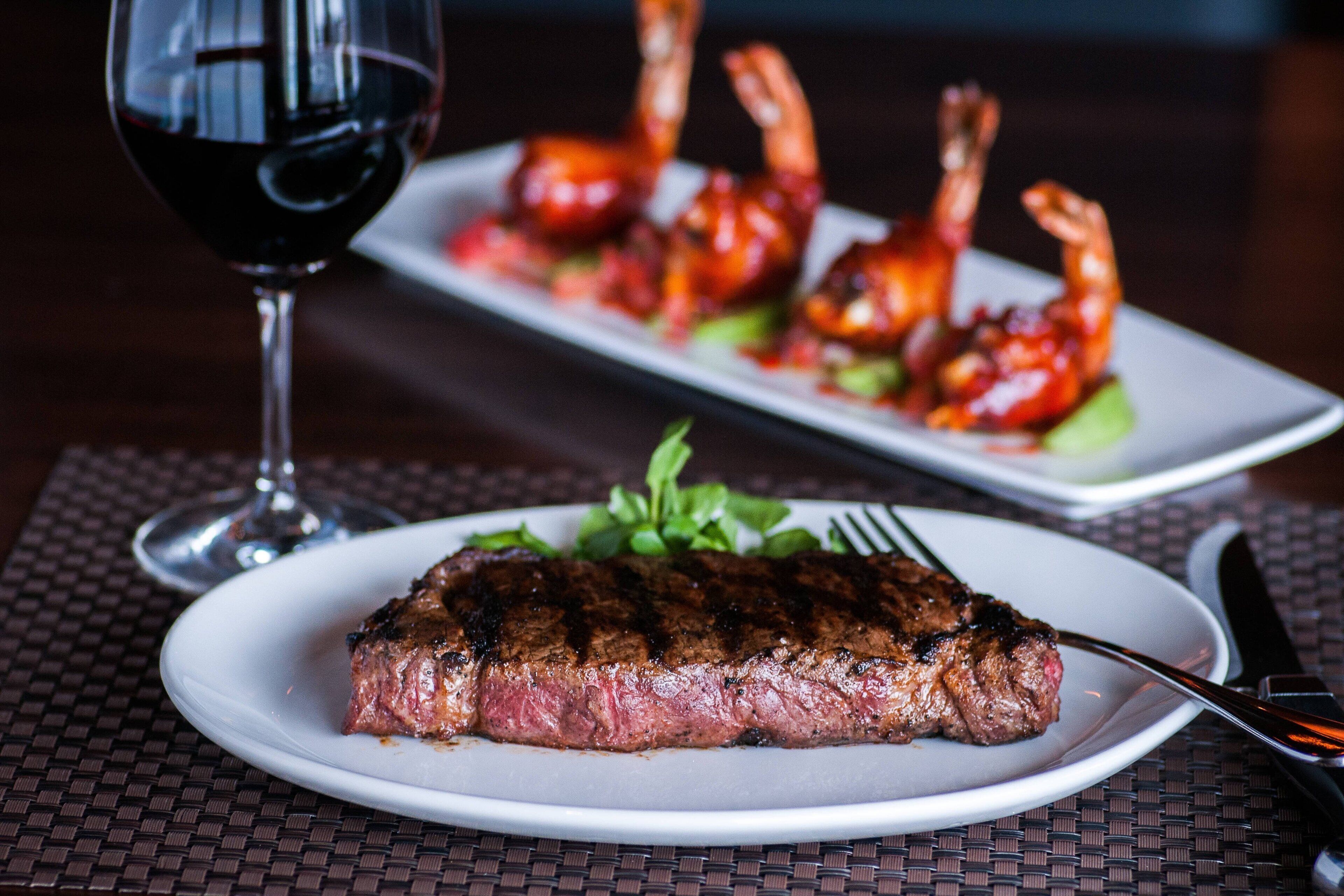 A steak with a side of vegetables and shrimp on a plate, a glass of red wine, and a fork and knife on a table.