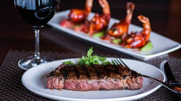 A steak with a side of vegetables and shrimp on a plate, a glass of red wine, and a fork and knife on a table.