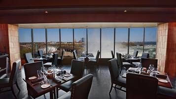 A restaurant with tables set for dining, large windows offering a view of a cityscape and a body of water.