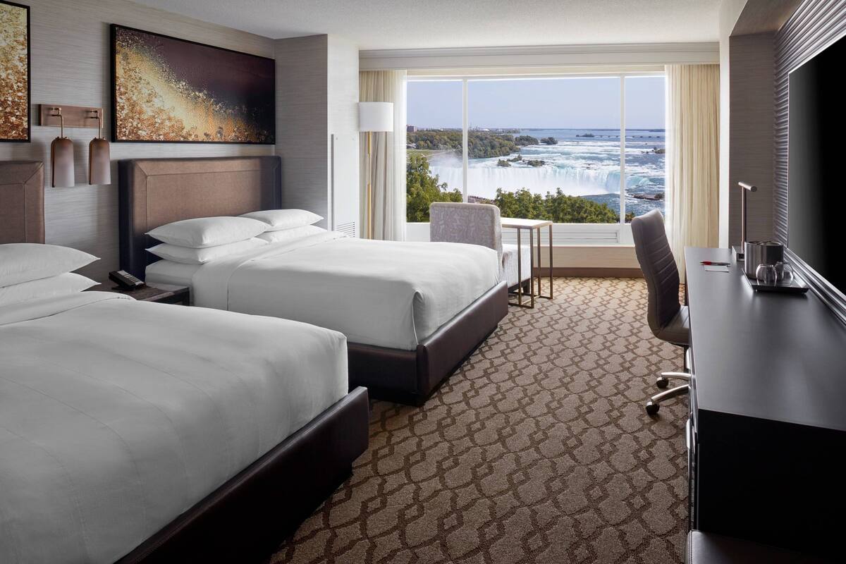 A hotel room with two beds, a desk, a chair, a TV, and a large window with a view of a waterfall.