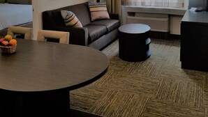 A hotel room with a bed, a sofa, a round table with fruit, a TV, and a window with curtains.