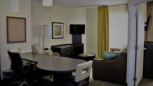 A hotel room with a dining table, two chairs, a sofa, a television, a desk, and a door.