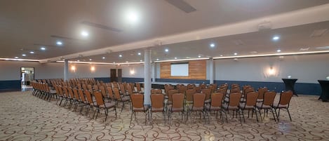 Un salón de conferencias con filas de sillas, una pantalla de proyector y una alfombra estampada.