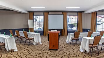 Un salón de conferencias con un atril, mesas blancas, sillas marrones y una pantalla de proyector.