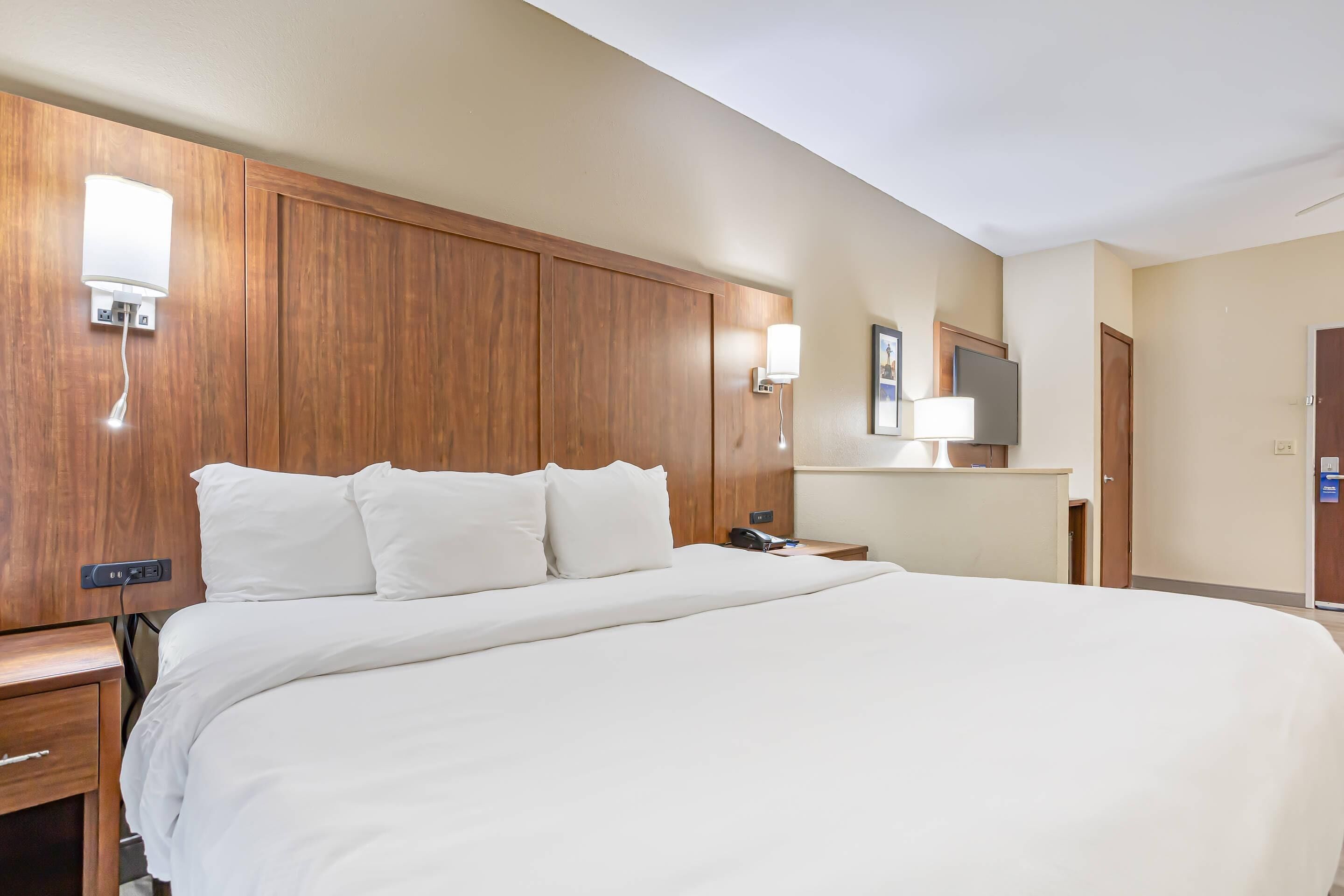 Une chambre d’hôtel avec un grand lit, une tête de lit en bois, des tables de chevet, une petite table, une lampe, un miroir et un téléviseur.