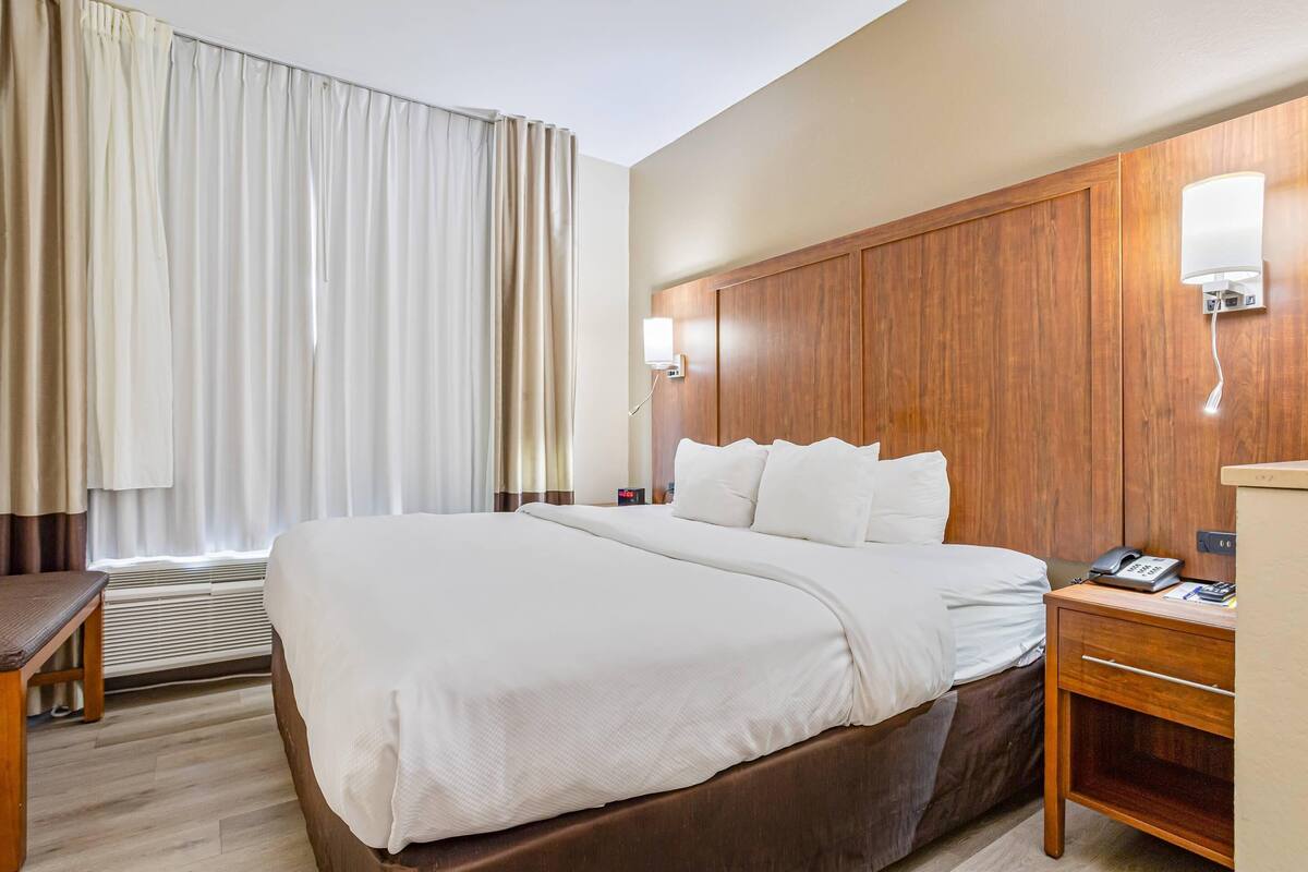 A hotel room with a large bed, wooden headboard, bedside table with a lamp, and a window with curtains.