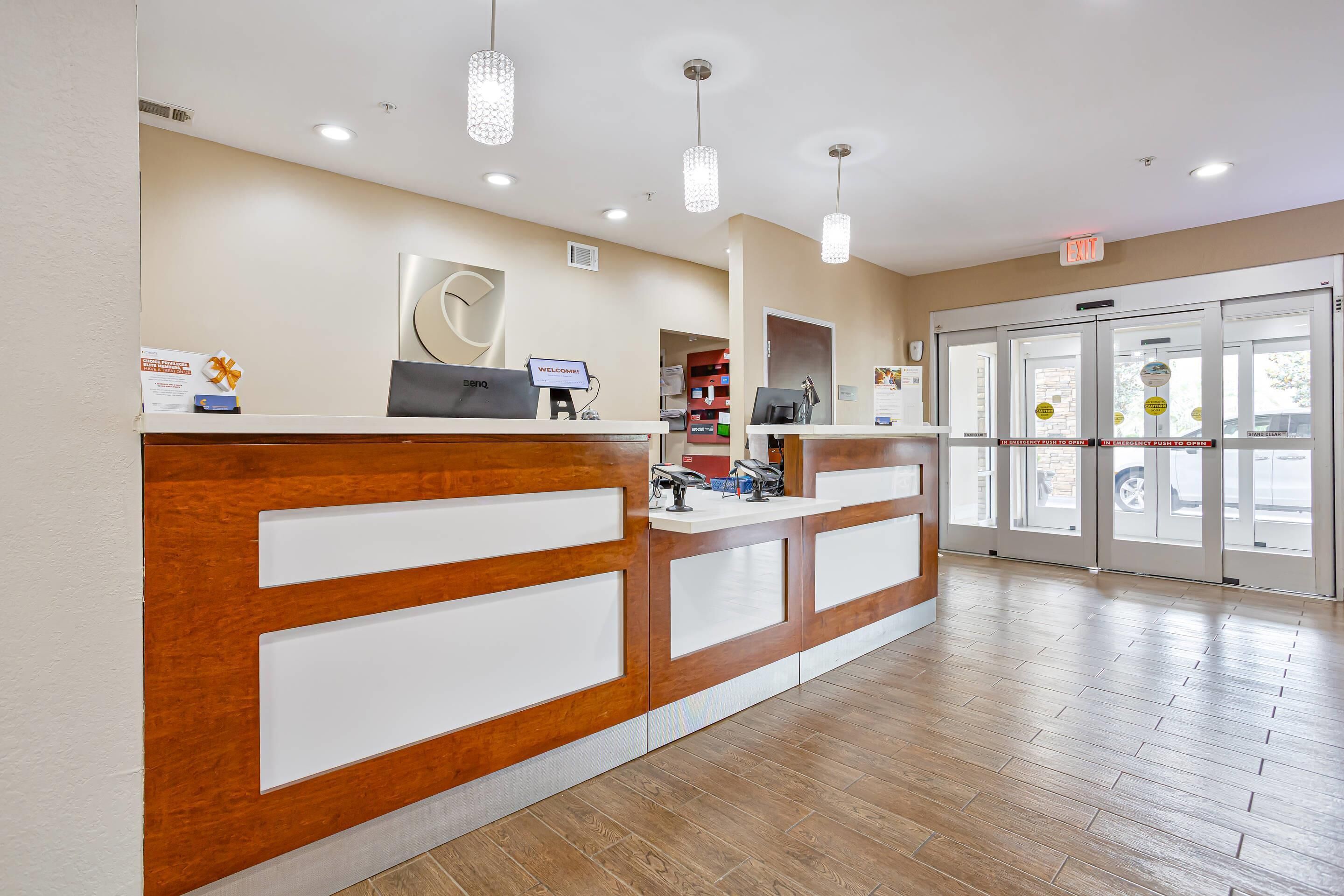A reception desk with a wooden finish and white panels, a computer, and a 'Welcome' sign. There are glass doors with a view outside, and a sign indicating an emergency exit.
