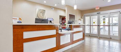 A reception desk with a wooden finish and white panels, a computer, and a 'Welcome' sign. There are glass doors with a view outside, and a sign indicating an emergency exit.