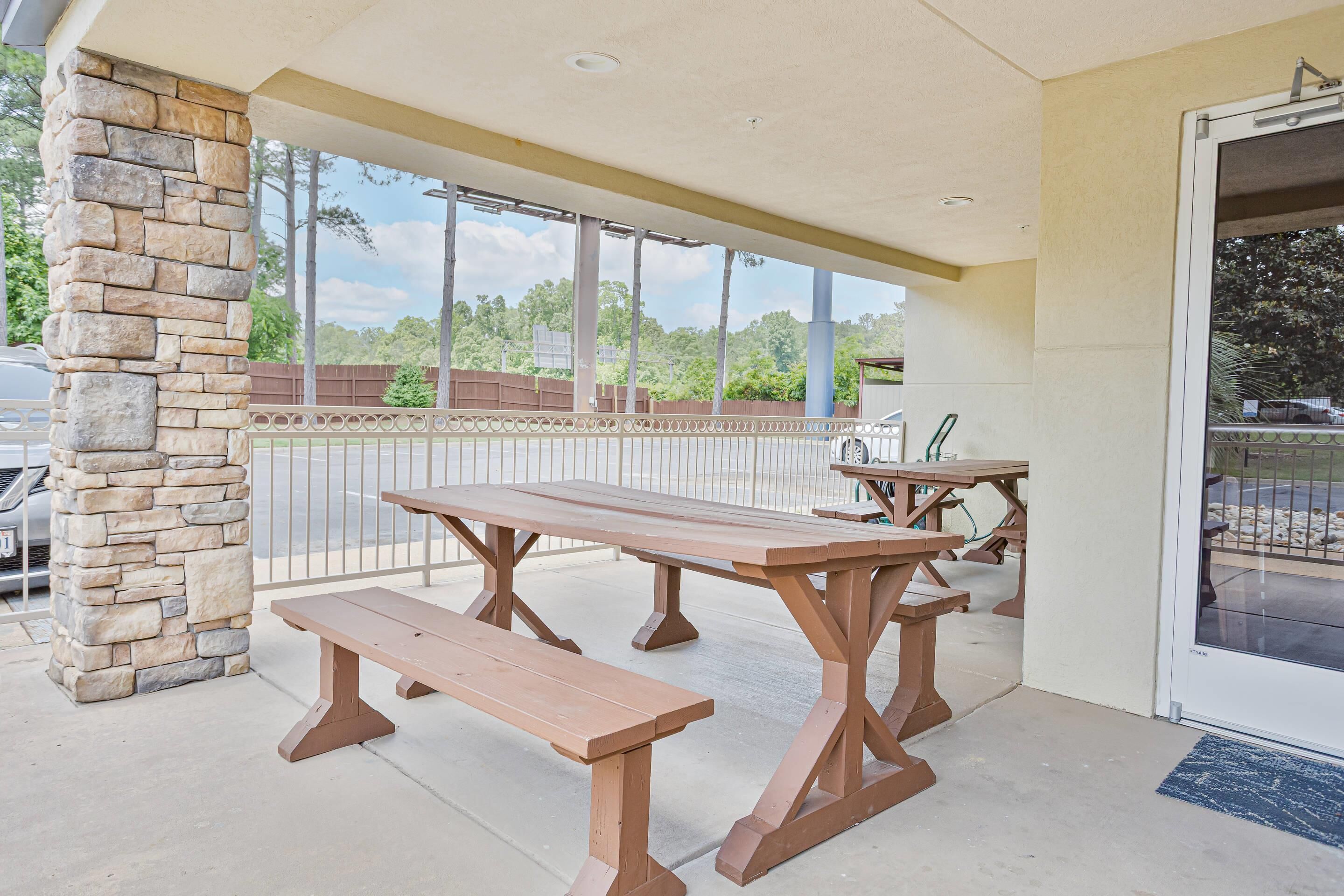 An outdoor patio with picnic tables and benches, a stone pillar, and a view of trees and a fence.