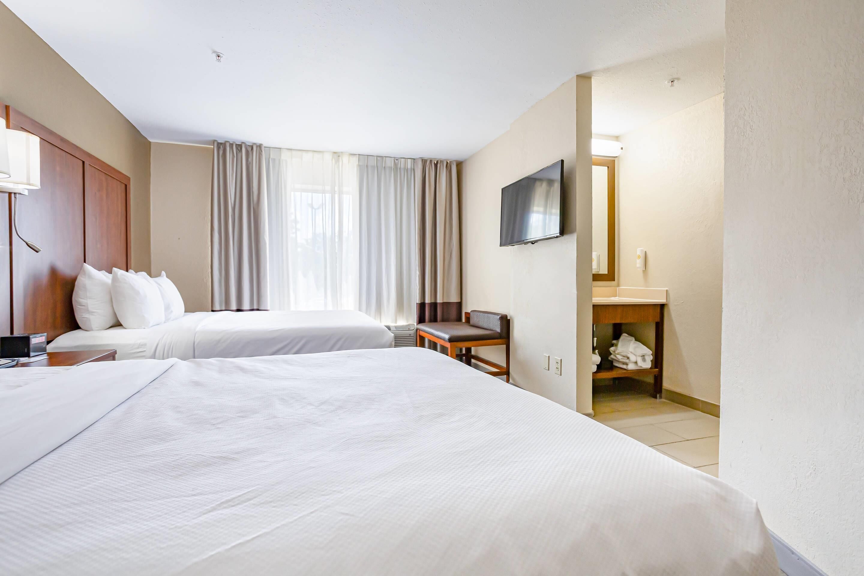 A hotel room with two beds, a desk, a TV, and a bathroom visible through an open door.