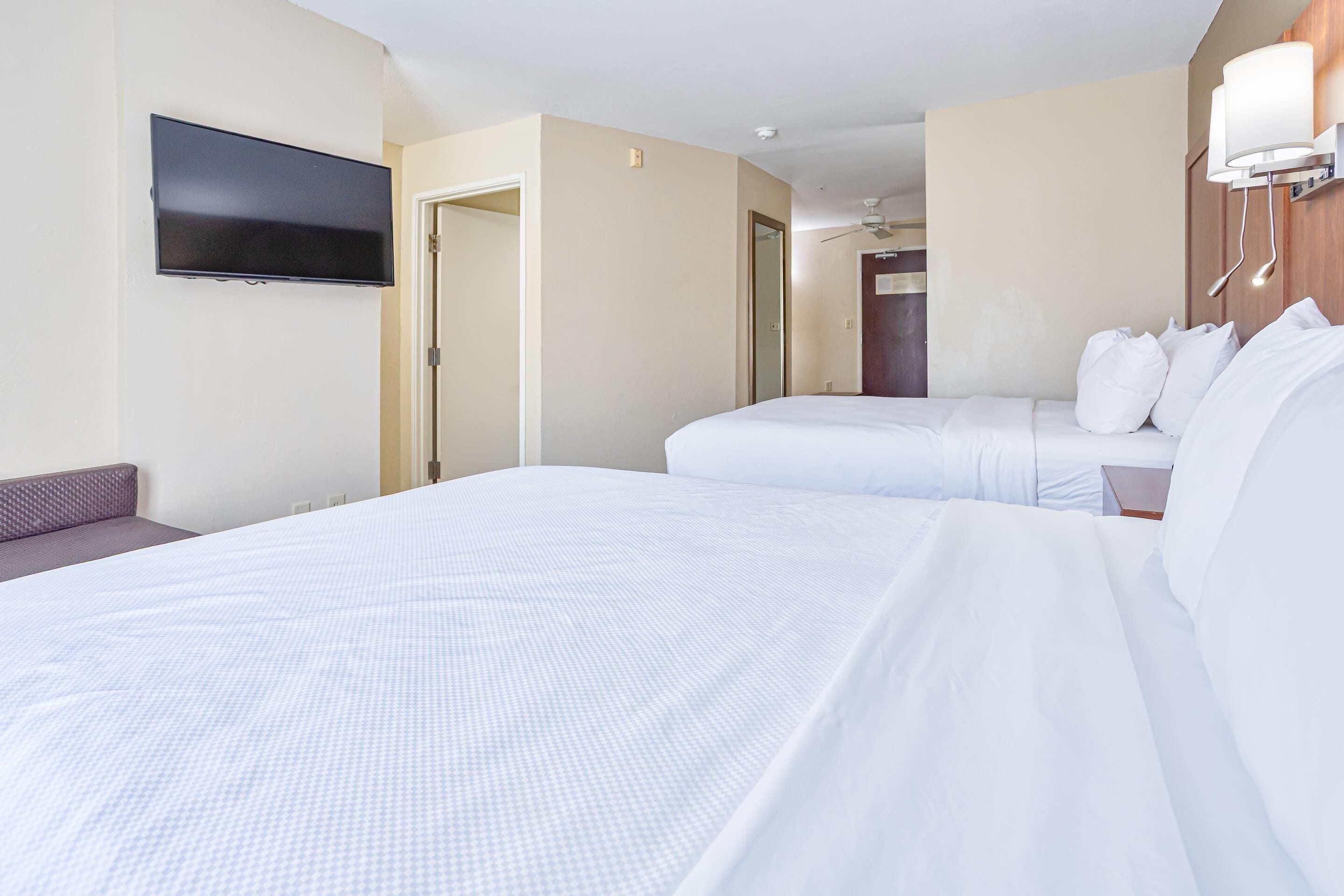 A hotel room with a large bed, a smaller bed, a flat-screen TV mounted on the wall, a ceiling fan, and a bedside table with a lamp.