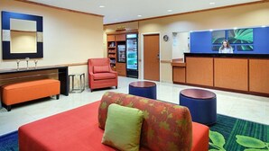 Un hall d’hôtel comprenant un comptoir d’accueil, un fauteuil rouge, un canapé agrémenté de coussins à motifs floraux et un réfrigérateur rempli de boissons.