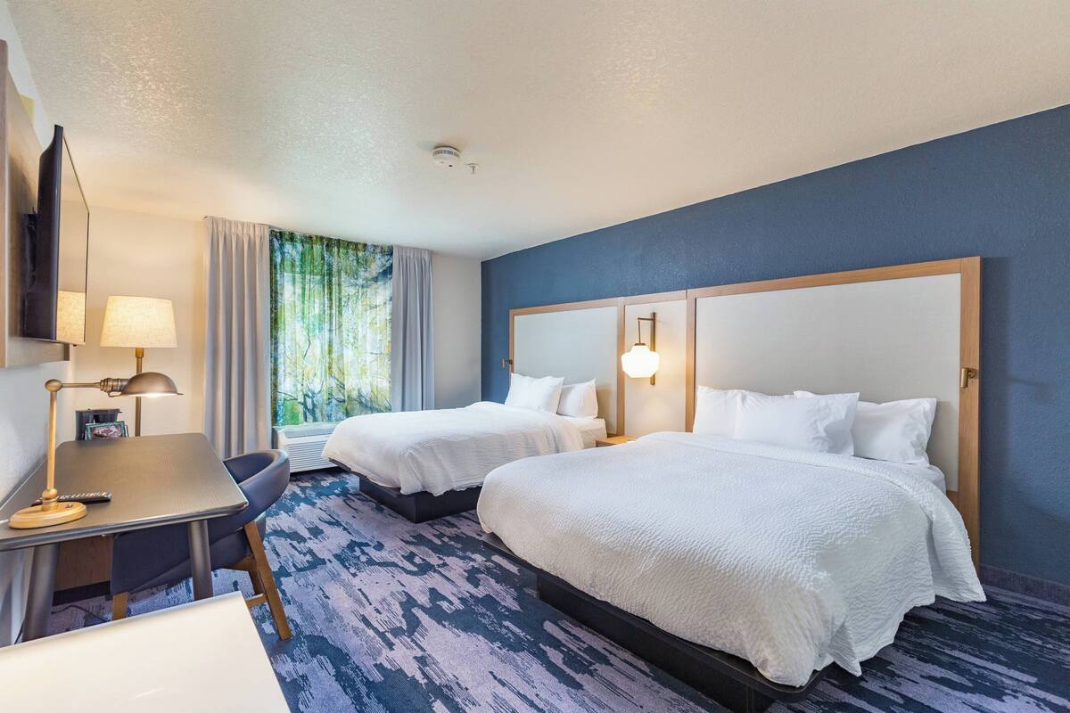 Ein Hotelzimmer mit zwei Betten, einem Schreibtisch, einem Stuhl, einer Lampe und einem Fenster mit Vorhängen.