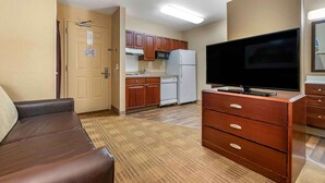 A compact living space with a kitchenette, a flat-screen TV, a brown leather sofa, and a wooden dresser.