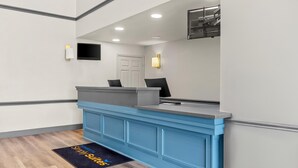 A reception desk with a blue counter, a flat-screen TV mounted on the wall, and a door in the background. - Sonesta Simply Suites Stafford (Stafford)