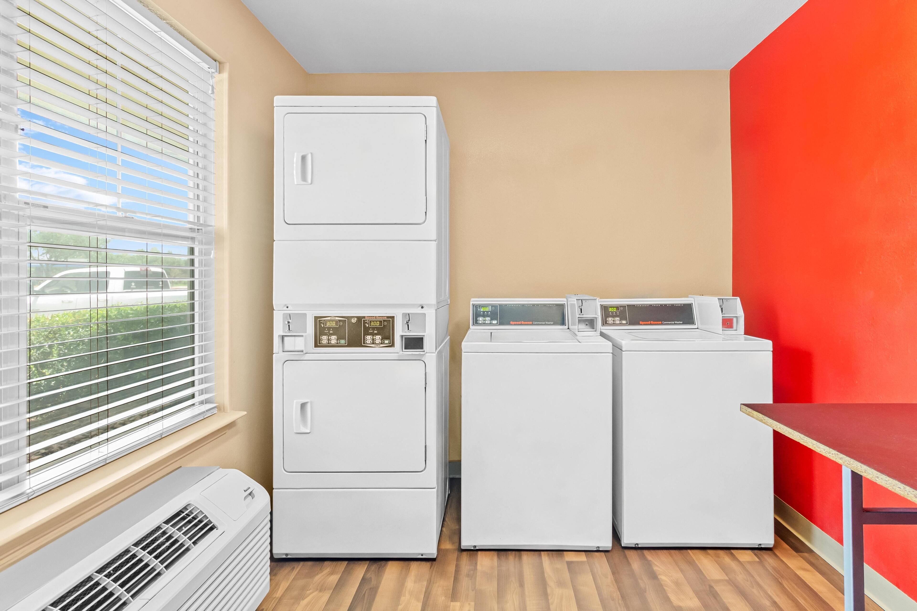 A laundry room with stacked washers and dryers, a table, and a window with blinds.