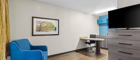 Une pièce avec une chaise bleue, un bureau, un téléviseur à écran plat, une commode et un tableau encadré accroché au mur.