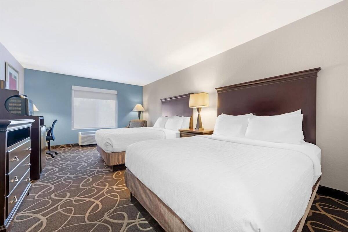 A hotel room with two beds, a desk, a chair, a lamp, and a window.