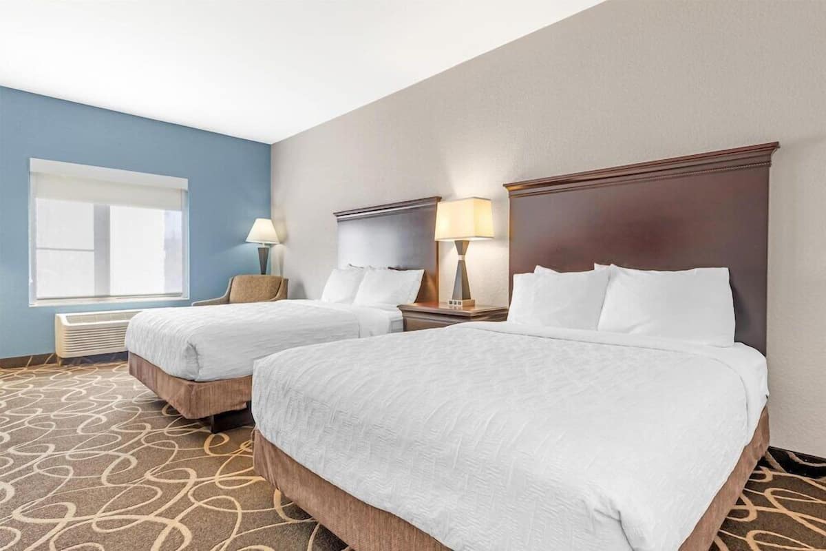 A hotel room with two beds, a window with blinds, a lamp, and a carpeted floor.