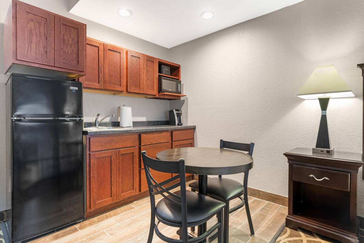 Una cocina compacta con armarios de madera, un refrigerador negro, un microondas, una mesa con sillas y una lámpara.