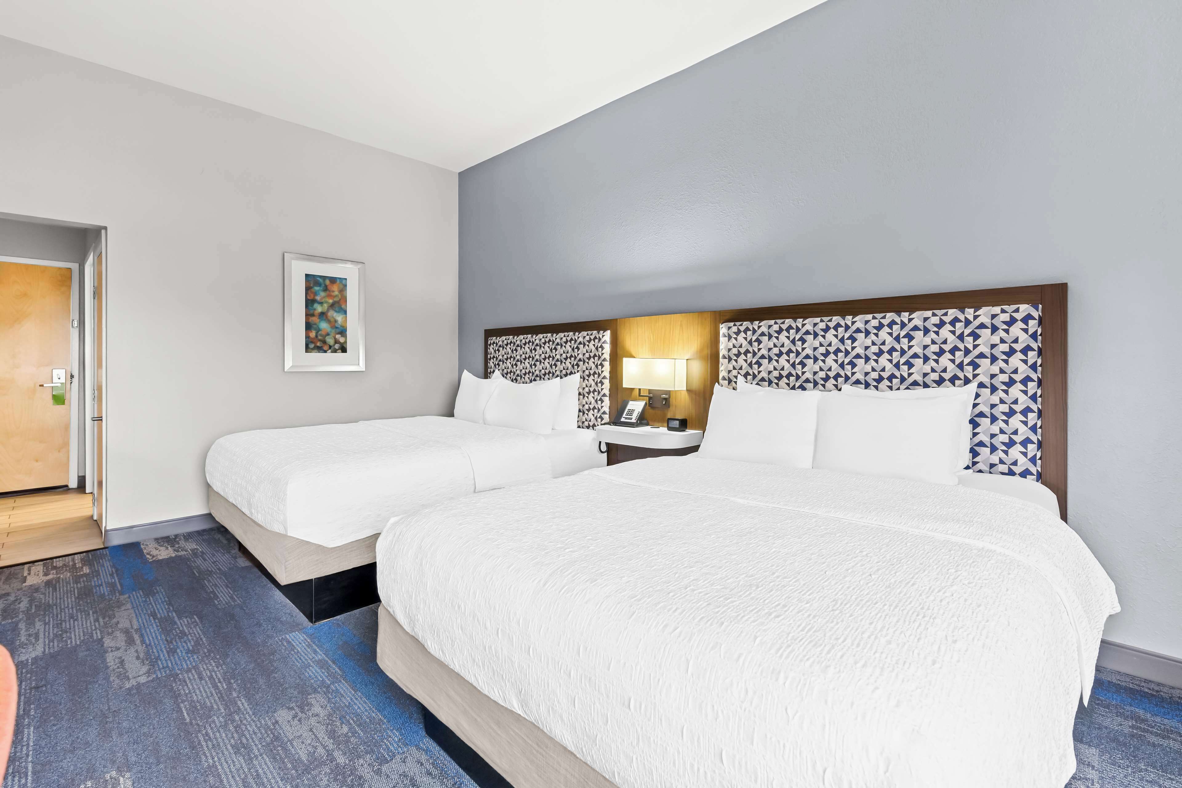 Quarto de hotel com duas camas, uma mesa de cabeceira, um abajur e um quadro na parede.