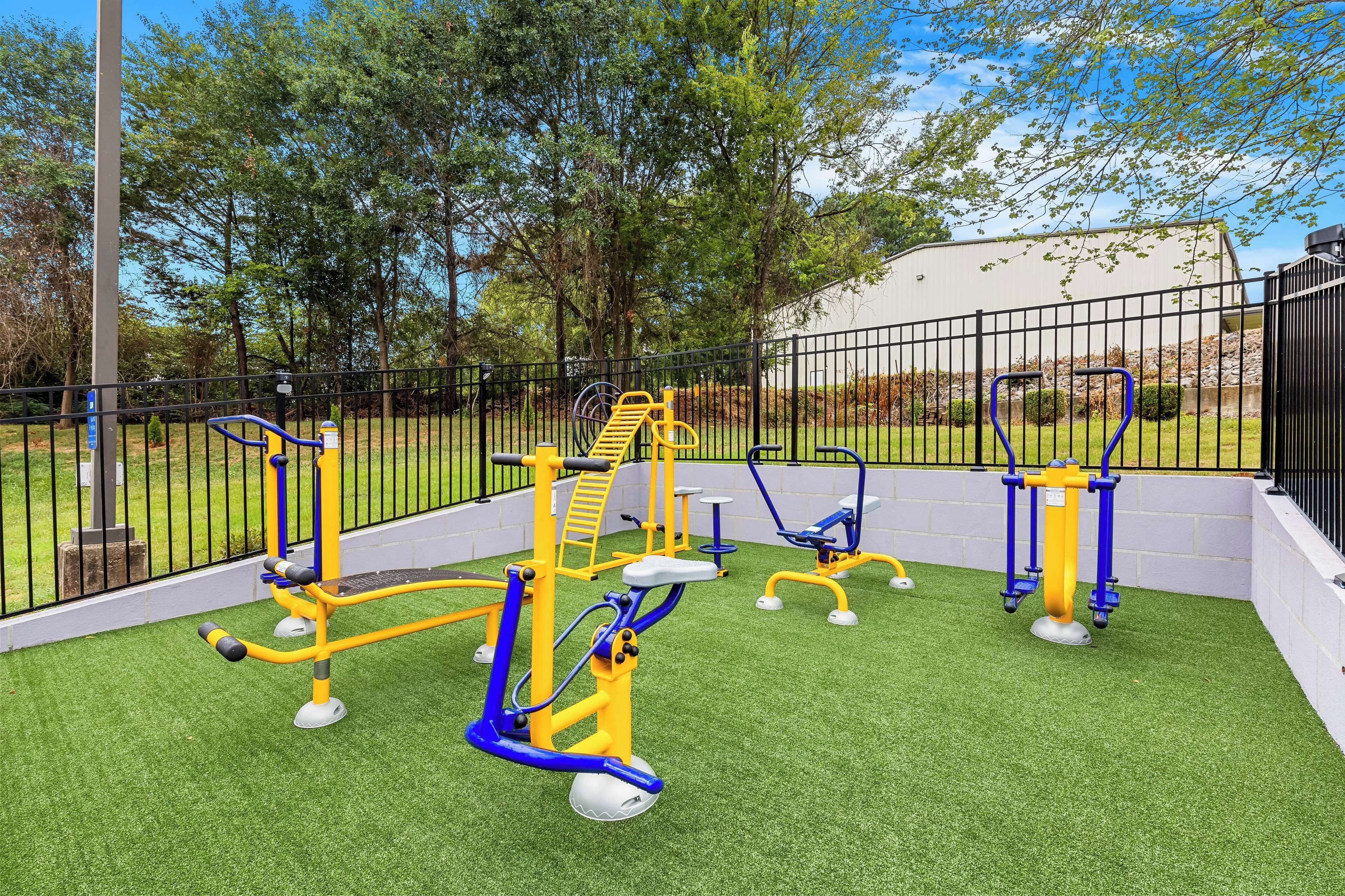 An outdoor fitness area with exercise equipment, surrounded by a fence and trees.