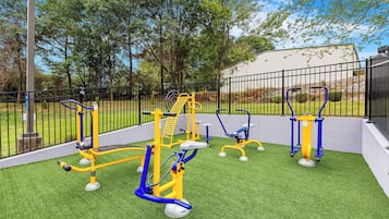 An outdoor fitness area with exercise equipment, surrounded by a fence and trees.