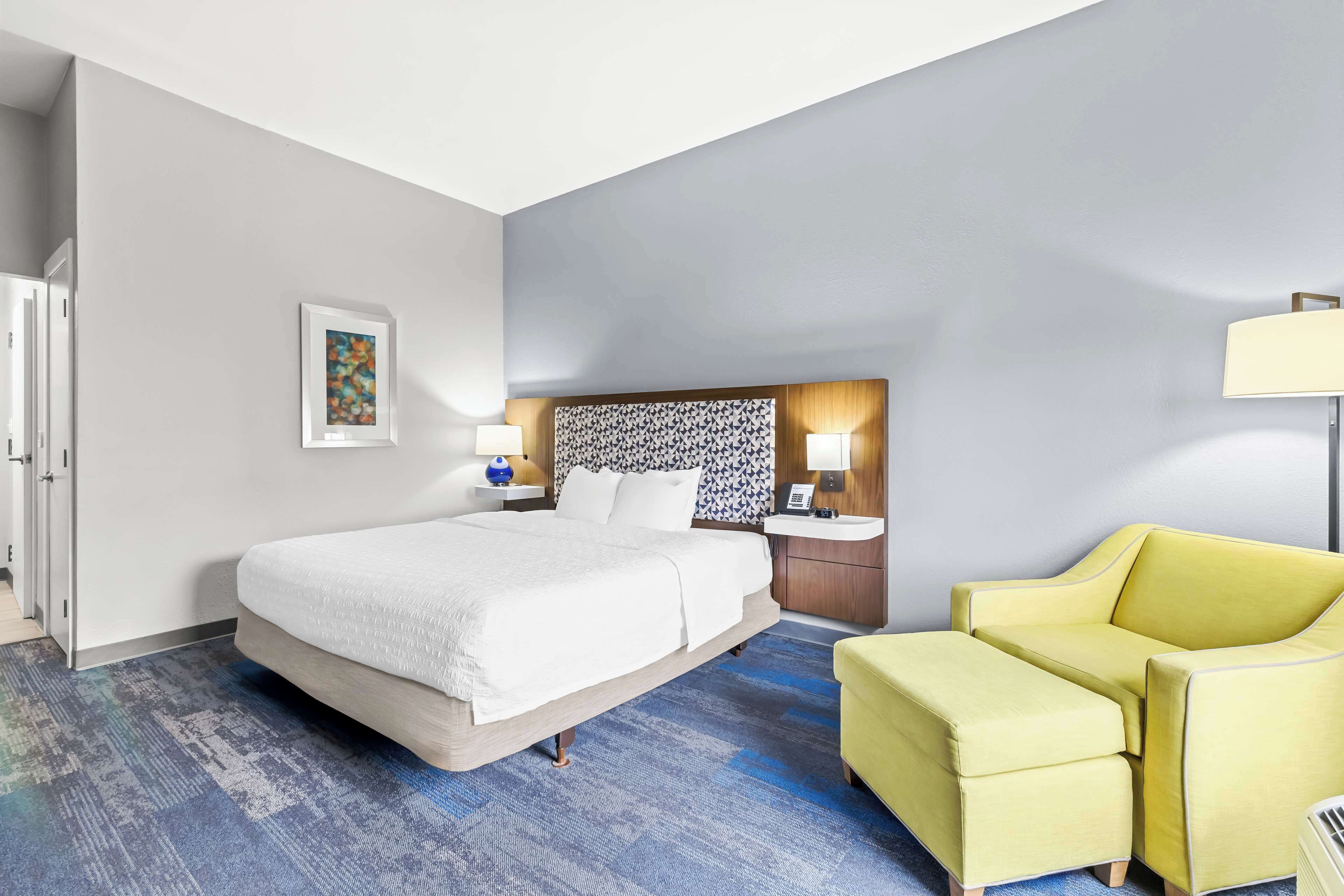 A hotel room with a large bed, a bedside table with lamps, a yellow armchair, and a headboard with a patterned design.
