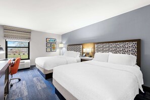 A hotel room with two beds, a desk, an orange chair, a window with a view, and a patterned headboard.