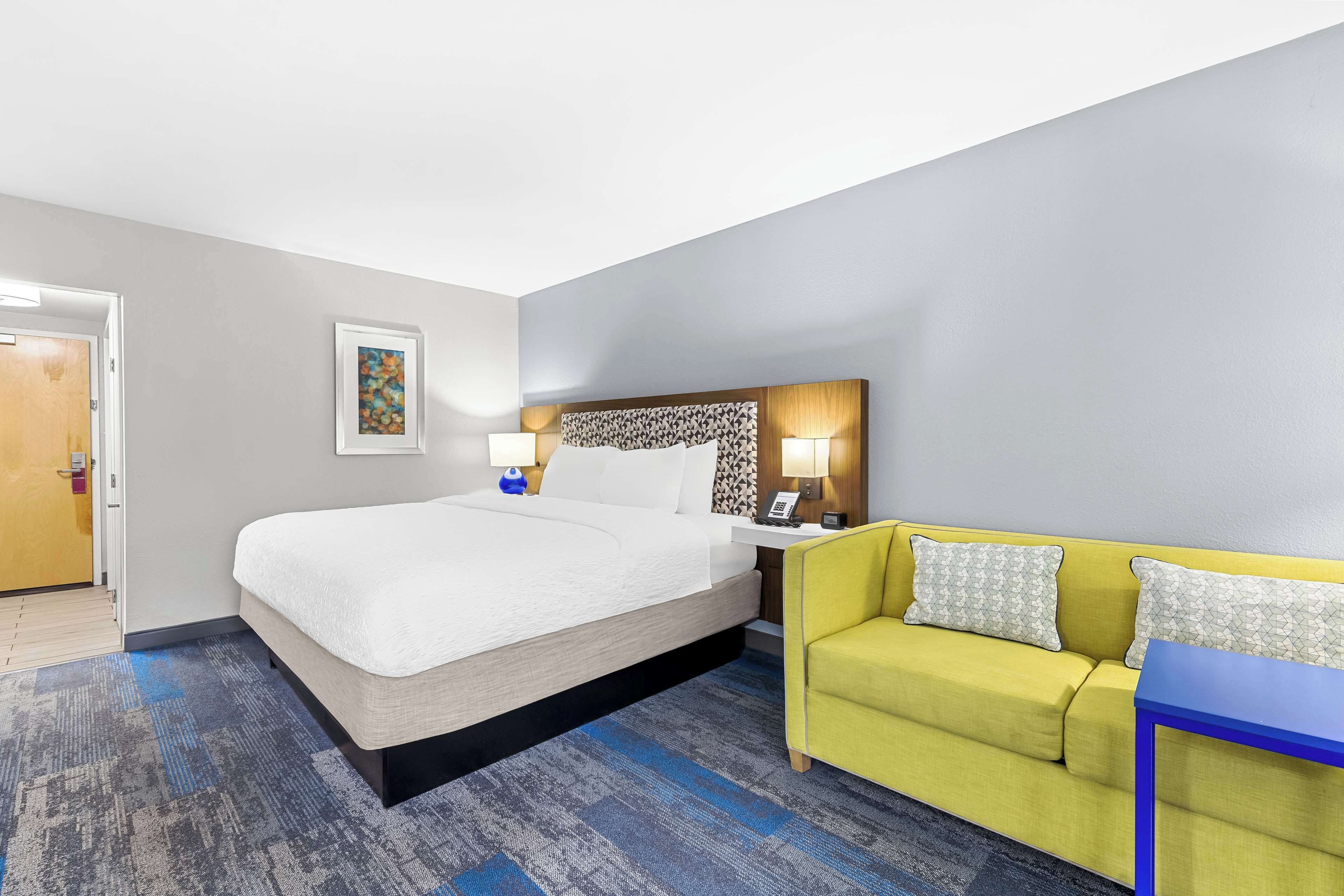 Une chambre d’hôtel avec un lit, un canapé jaune, une table bleue et une tête de lit ornée d’un motif.