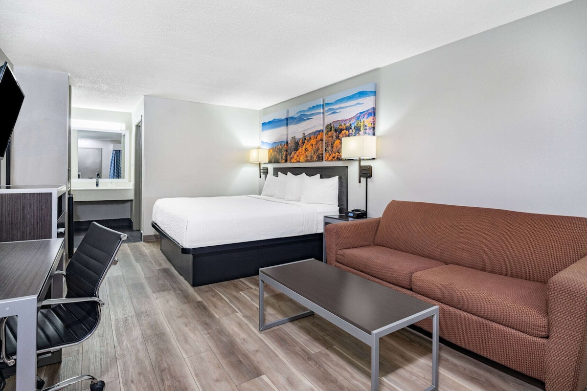 A hotel room with a bed, a brown sofa, a small table, a desk with a chair, and a painting on the wall.