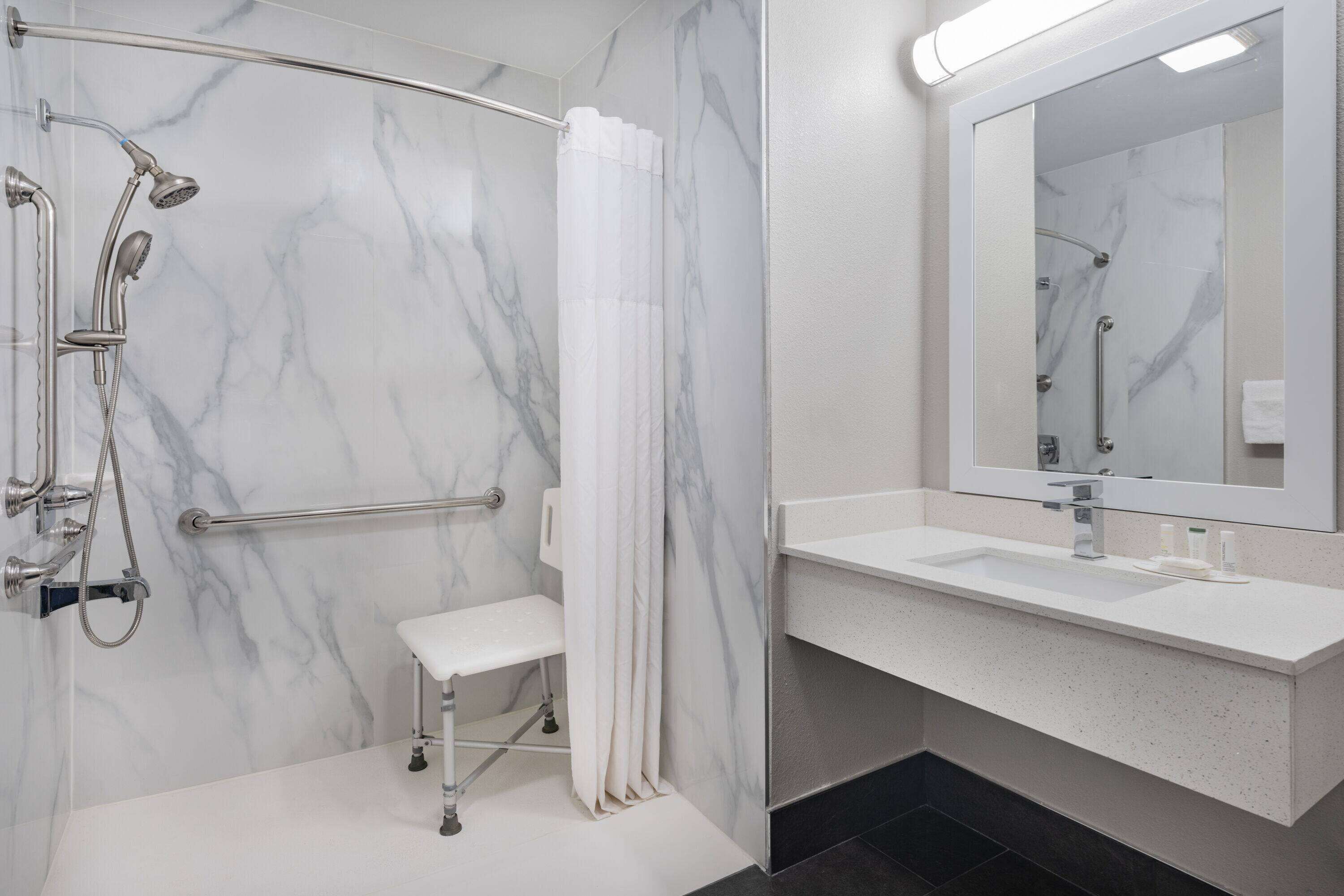 Un baño moderno con ducha a nivel, un banco, un espejo grande y un lavamanos blanco.