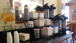 Um balcão de café da manhã com diversos recipientes de comida e bebida, incluindo um dispensador de ponche de frutas, máquinas de café e suportes para guardanapos.