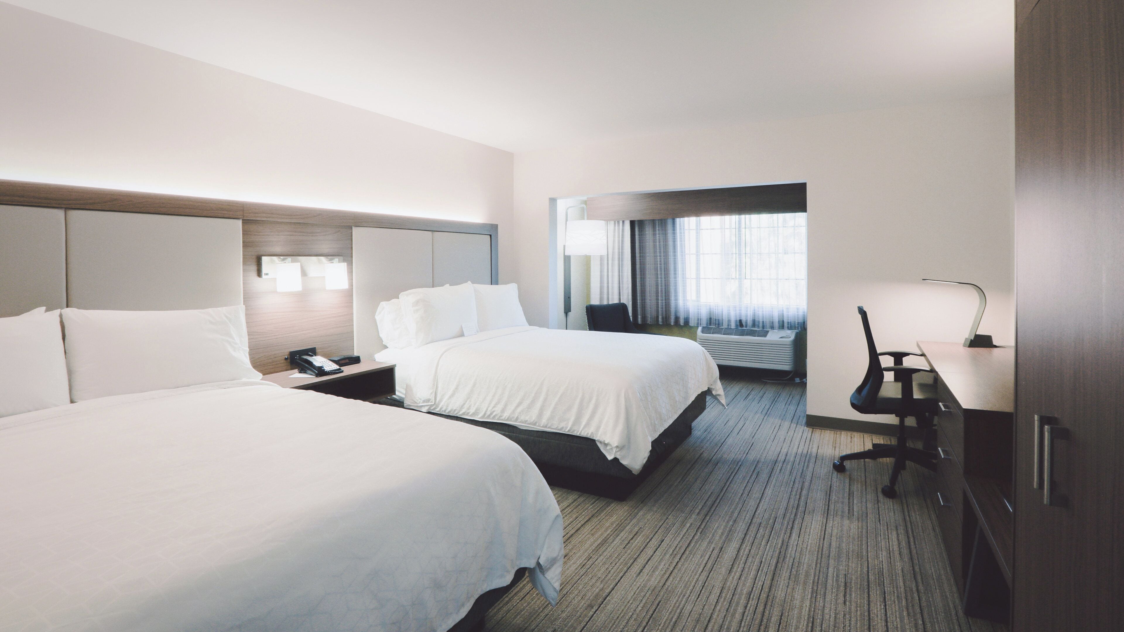 A hotel room with two beds, a desk, a chair, a lamp, and a window with curtains.