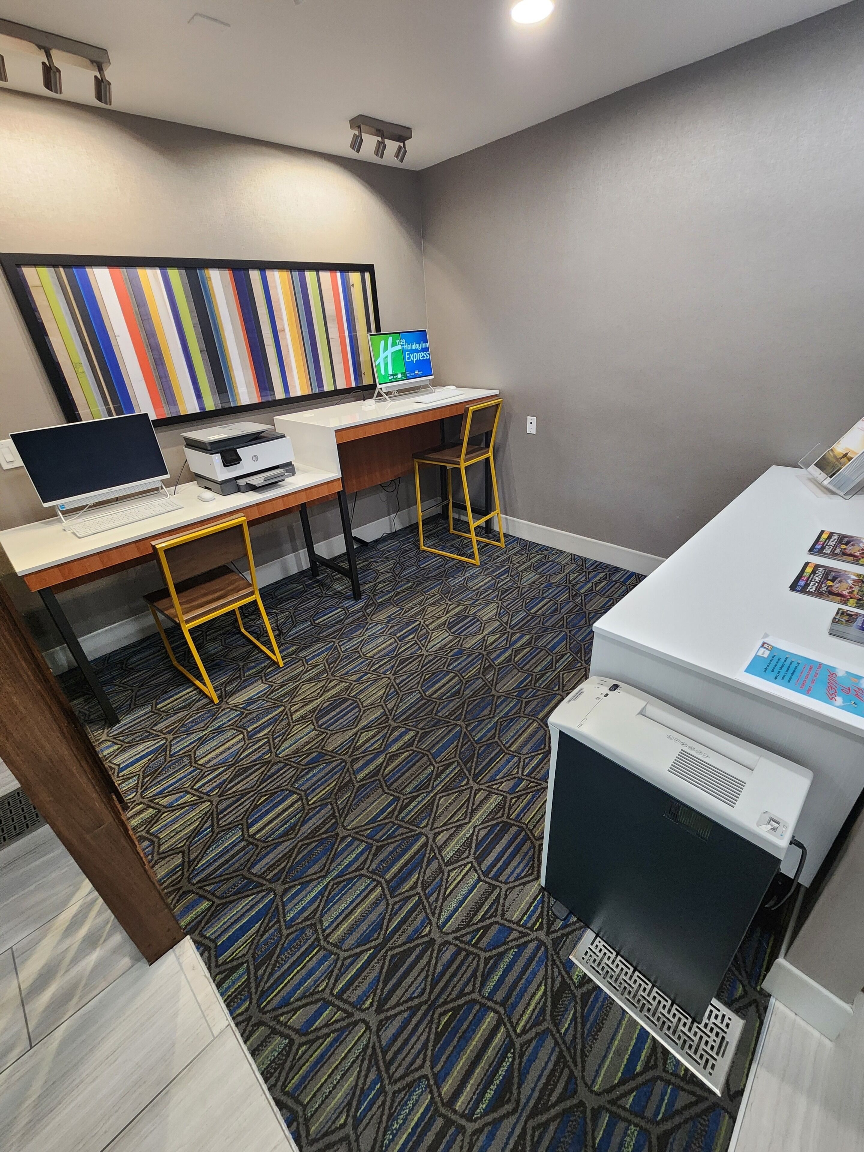 Quarto de hotel com escrivaninha, computador, impressora e uma obra de arte colorida listrada na parede.