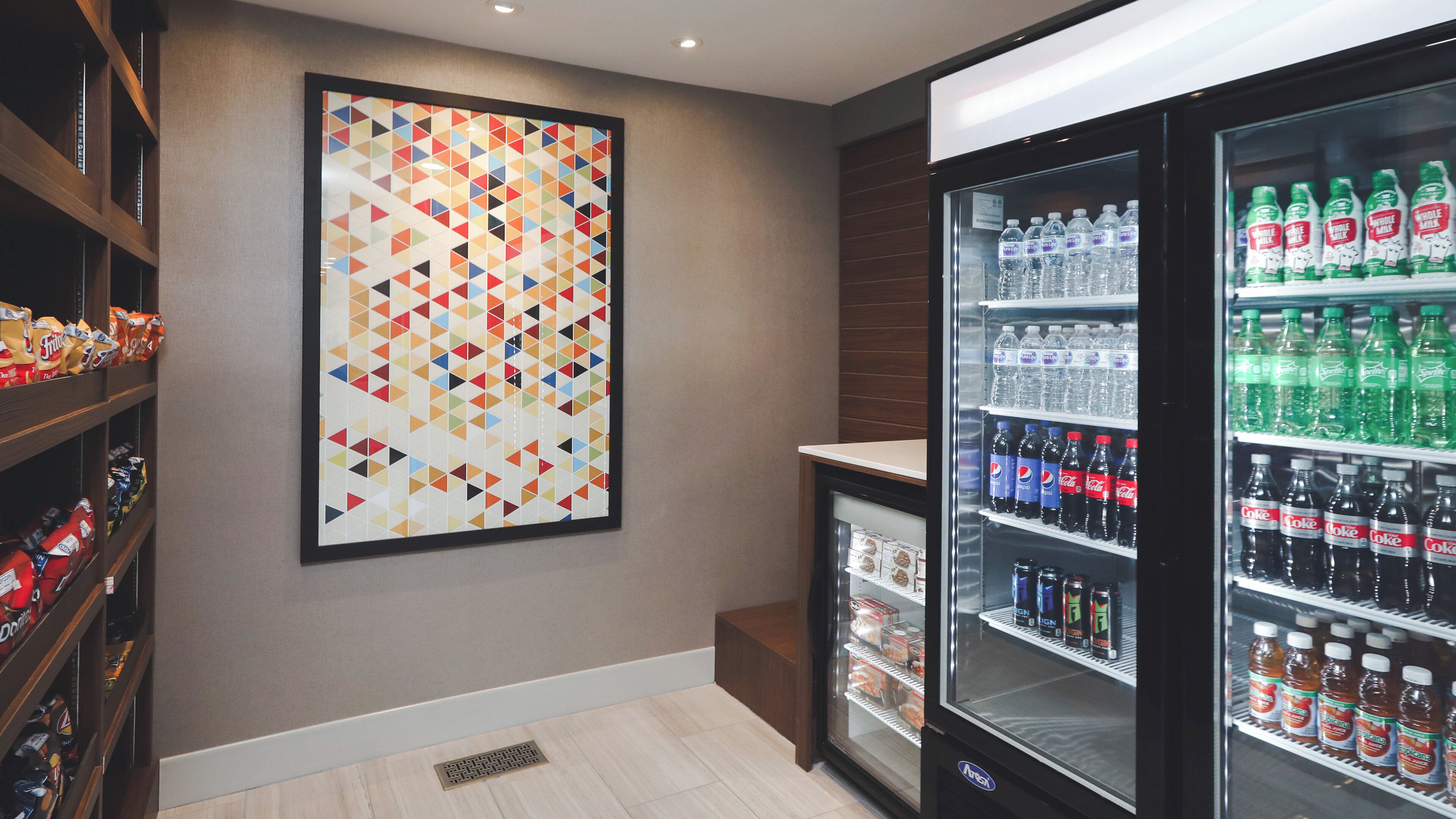A hotel room with a large geometric artwork on the wall, a mini-fridge stocked with beverages, and shelves with snacks.