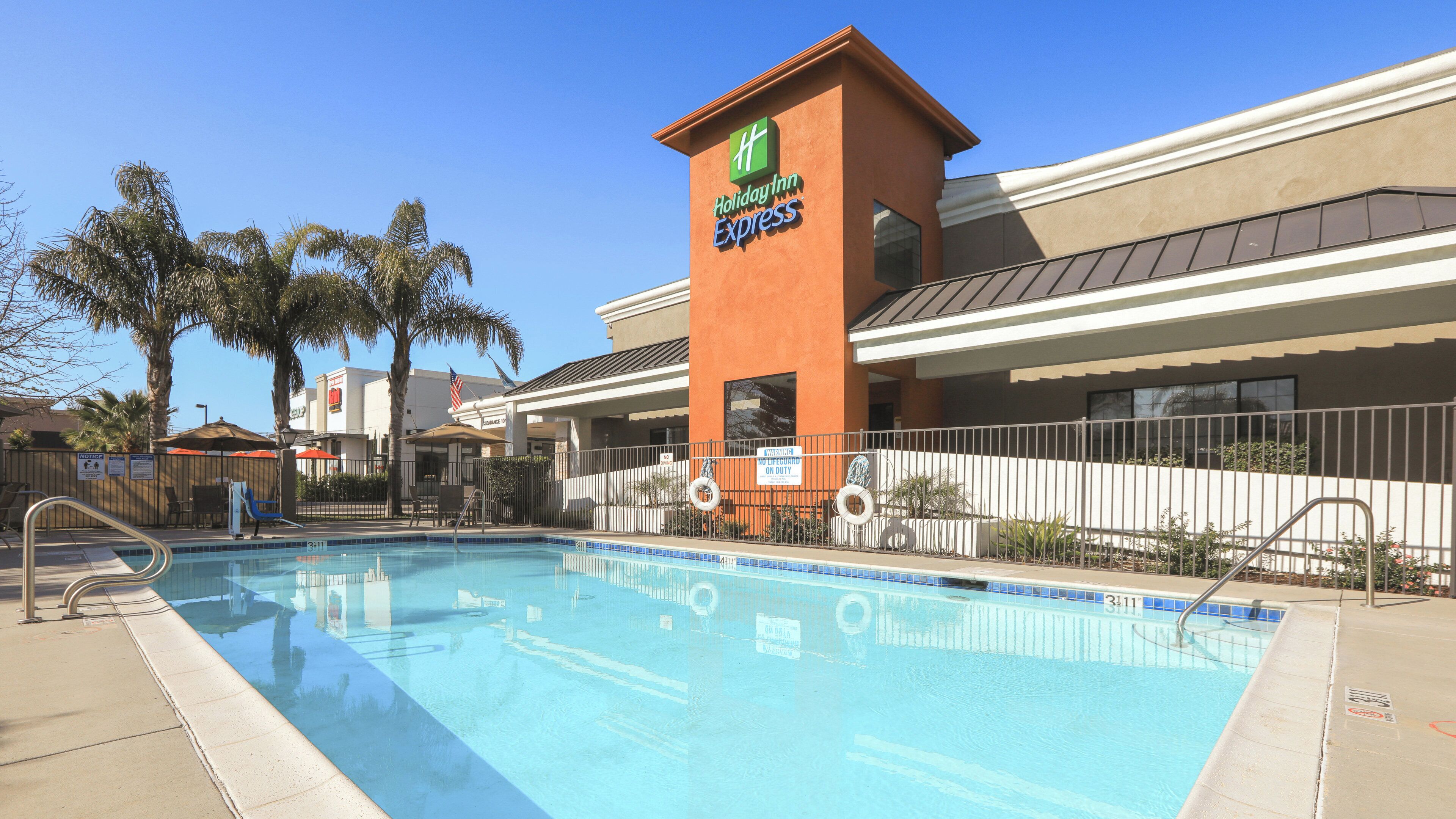 Ein Holiday Inn Express mit Schwimmbad, Palmen und einem eingezäunten Bereich.