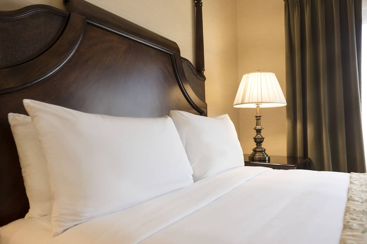 Una cama bien hecha con sábanas blancas, cabecera de madera y mesita de noche con lámpara.