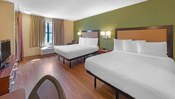 A hotel room with two beds, a desk with a computer, a TV, and a window with curtains. - Extended Hotel Studio (San Dimas)